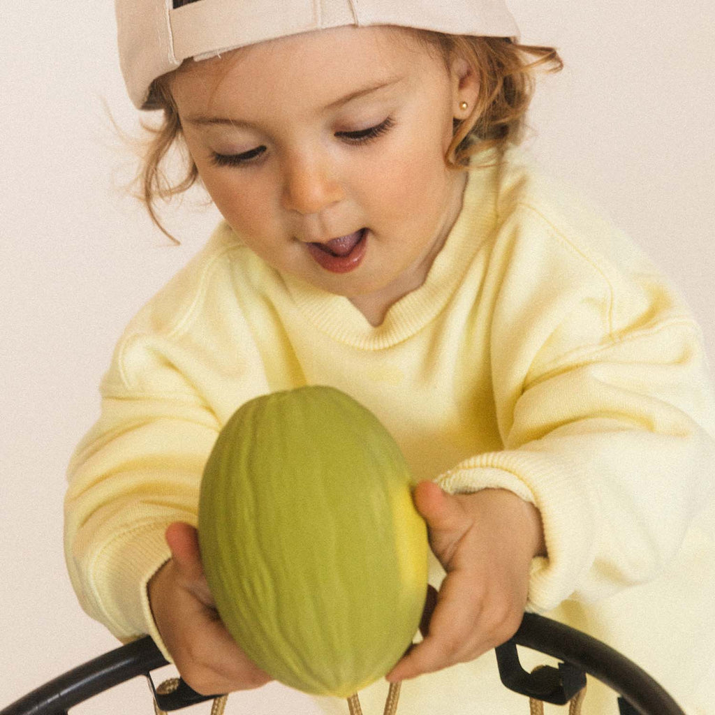 A young child in a yellow sweater and beige cap eagerly examines a green toy fruit while leaning over a black wire basket. The mood is playful and curious.