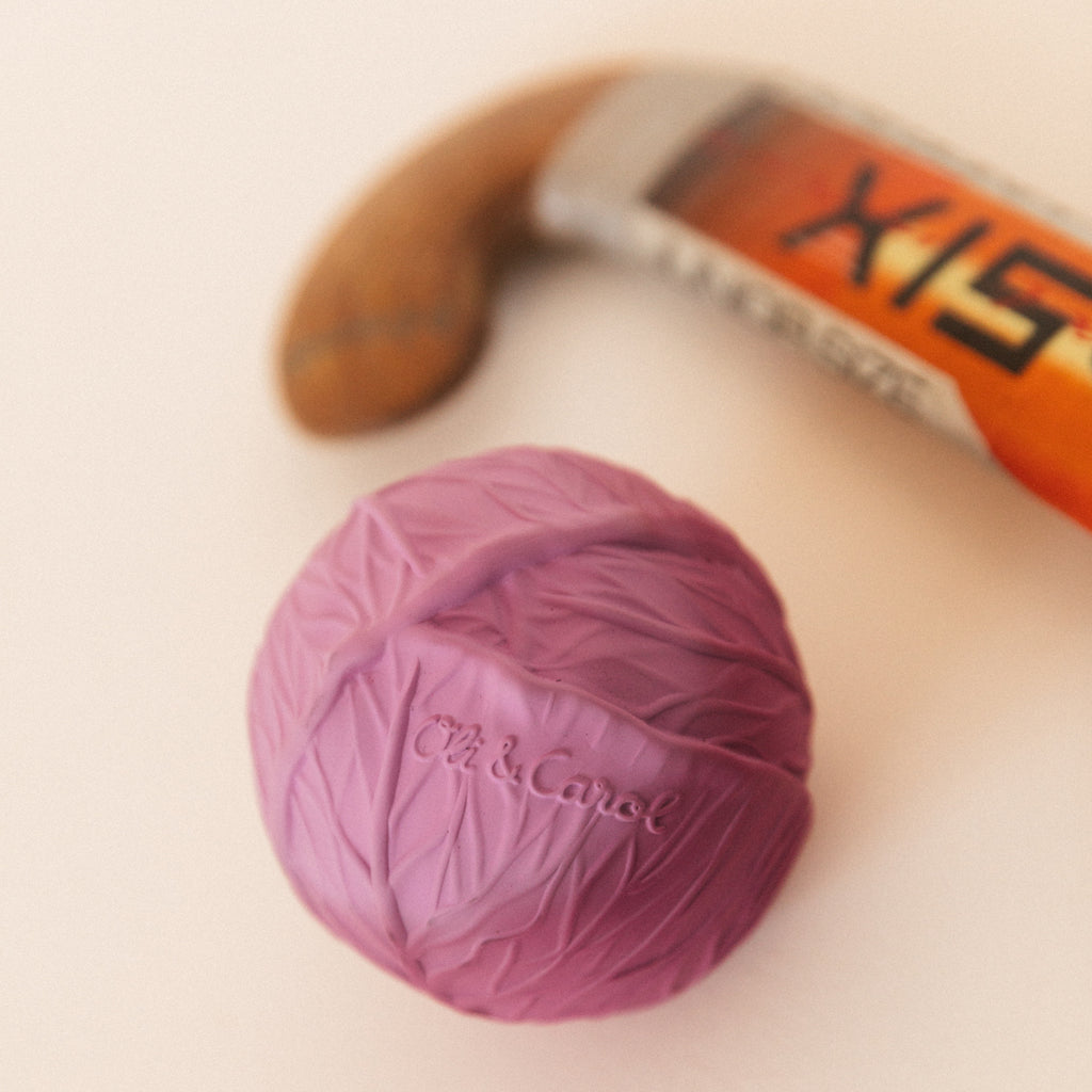 Purple cabbage-shaped toy with "Oli & Carol" embossed on it, beside a vintage hockey stick with a wooden handle and a partially visible orange head.
