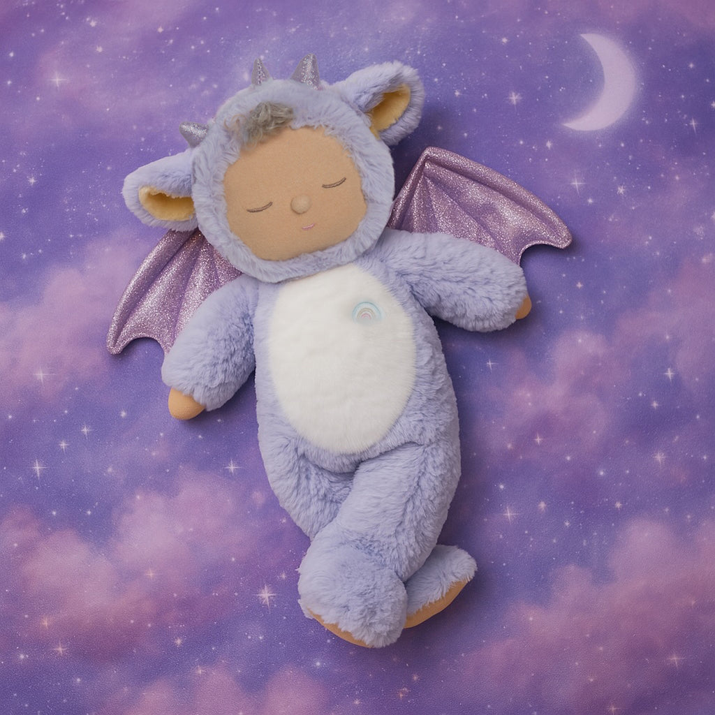 Cute plush doll in a purple dragon costume with sparkly wings. The doll has a serene expression, curly hair, and a small rainbow emblem.