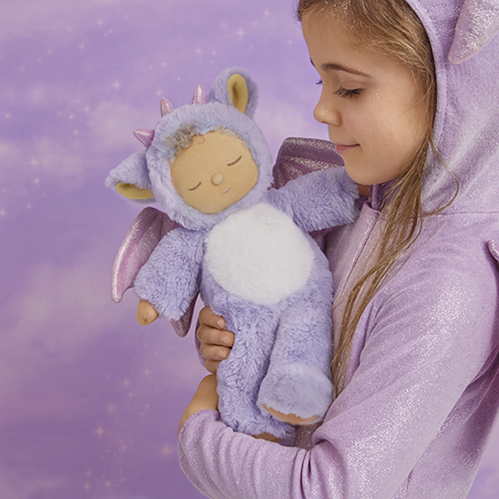 Cute plush doll in a purple dragon costume with sparkly wings. The doll has a serene expression, curly hair, and a small rainbow emblem.