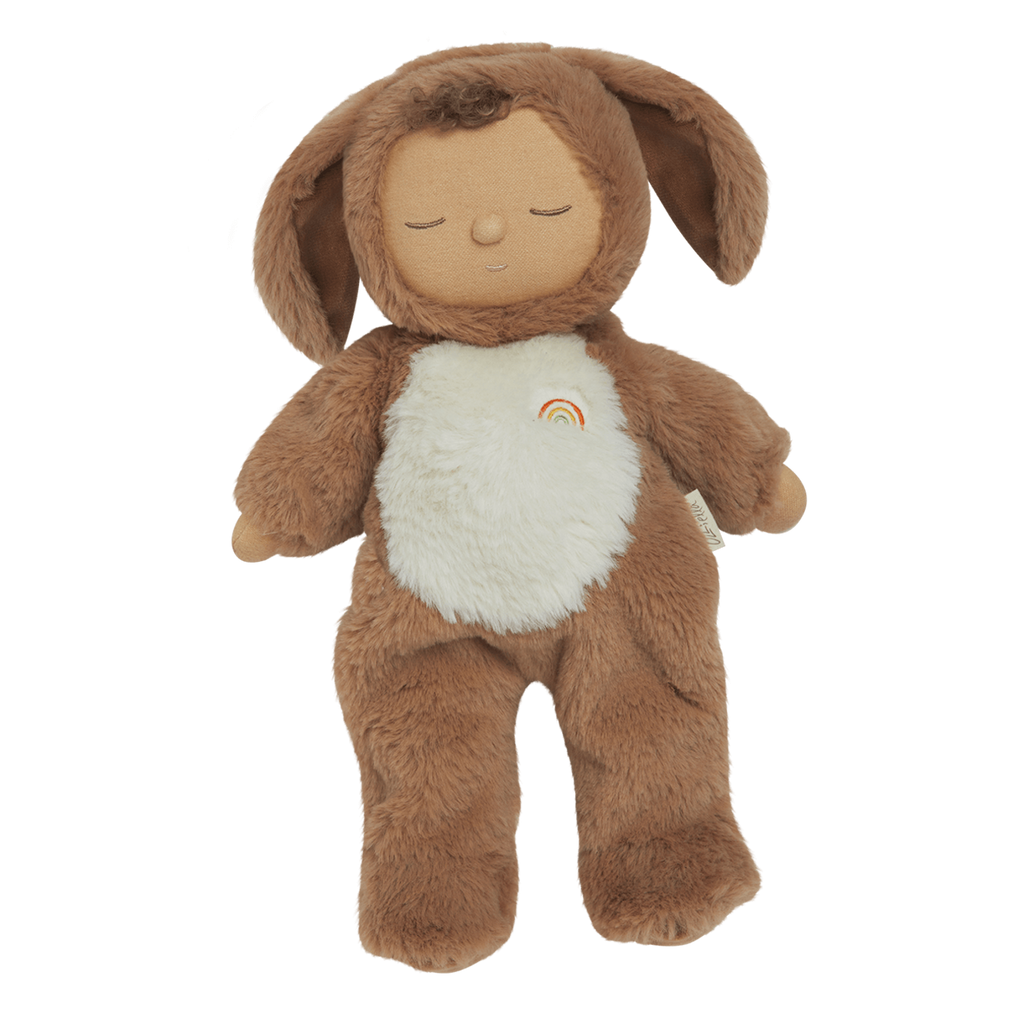 A plush toy with a brown, fluffy dog feature, closed eyes, and a serene expression. It features a white belly with a small embroidered rainbow.