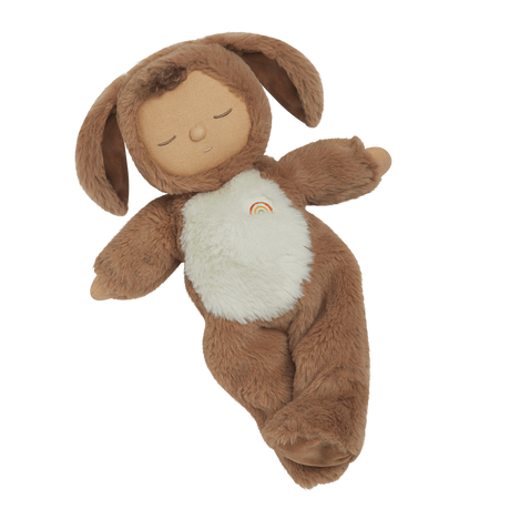 A plush toy with a brown, fluffy dog feature, closed eyes, and a serene expression. It features a white belly with a small embroidered rainbow.