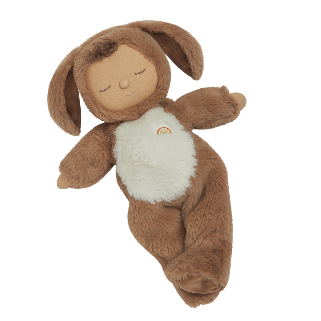 A plush toy with a brown, fluffy dog feature, closed eyes, and a serene expression. It features a white belly with a small embroidered rainbow.