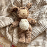 A plush toy with a brown, fluffy dog feature, closed eyes, and a serene expression. It features a white belly with a small embroidered rainbow.