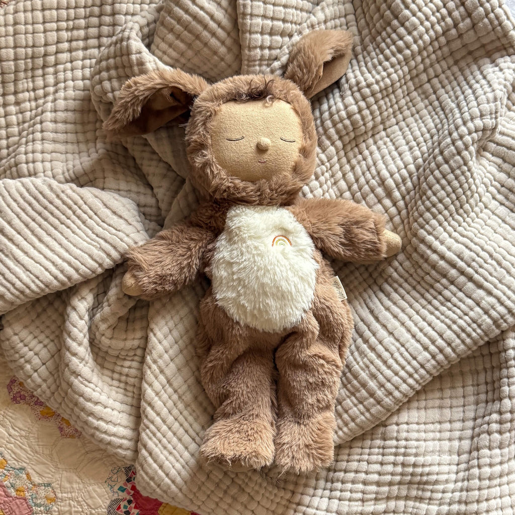 A plush toy with a brown, fluffy dog feature, closed eyes, and a serene expression. It features a white belly with a small embroidered rainbow.