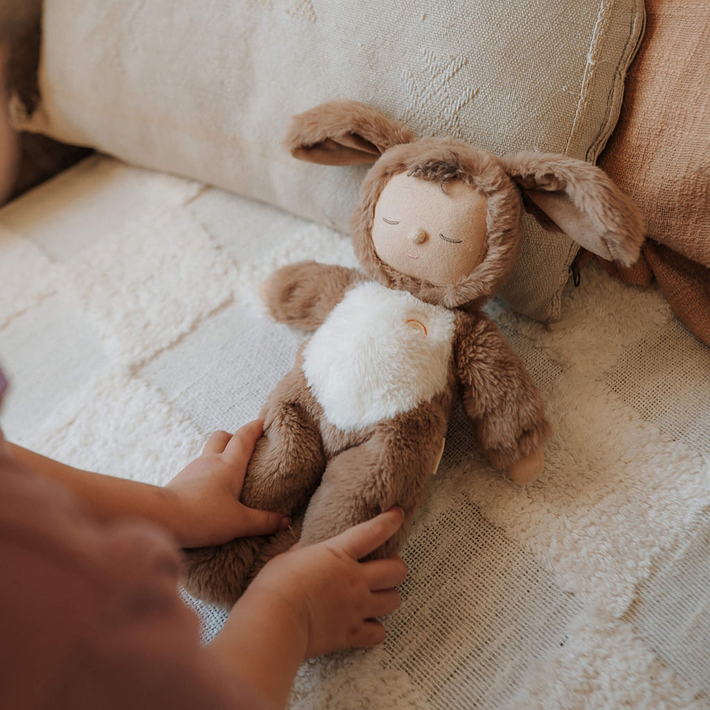 A plush toy with a brown, fluffy dog feature, closed eyes, and a serene expression. It features a white belly with a small embroidered rainbow.