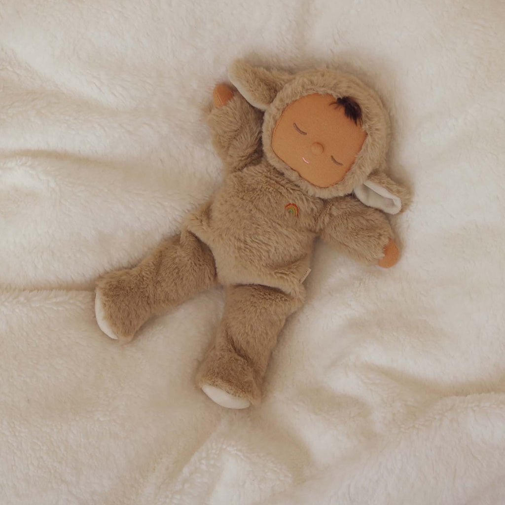 Cute doll in a fluffy beige sheep costume with floppy ears and a serene face. The doll wears a small embroidered rainbow, conveying calmness.