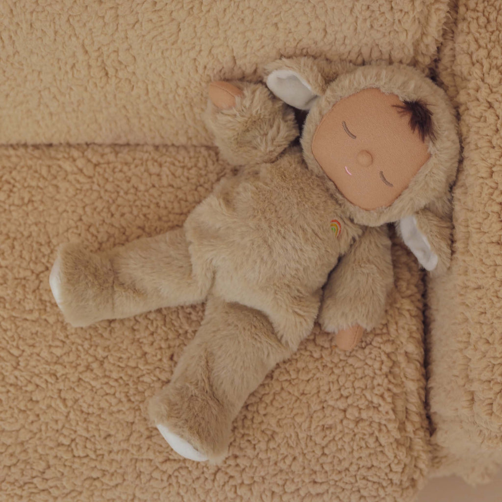Cute doll in a fluffy beige sheep costume with floppy ears and a serene face. The doll wears a small embroidered rainbow, conveying calmness.