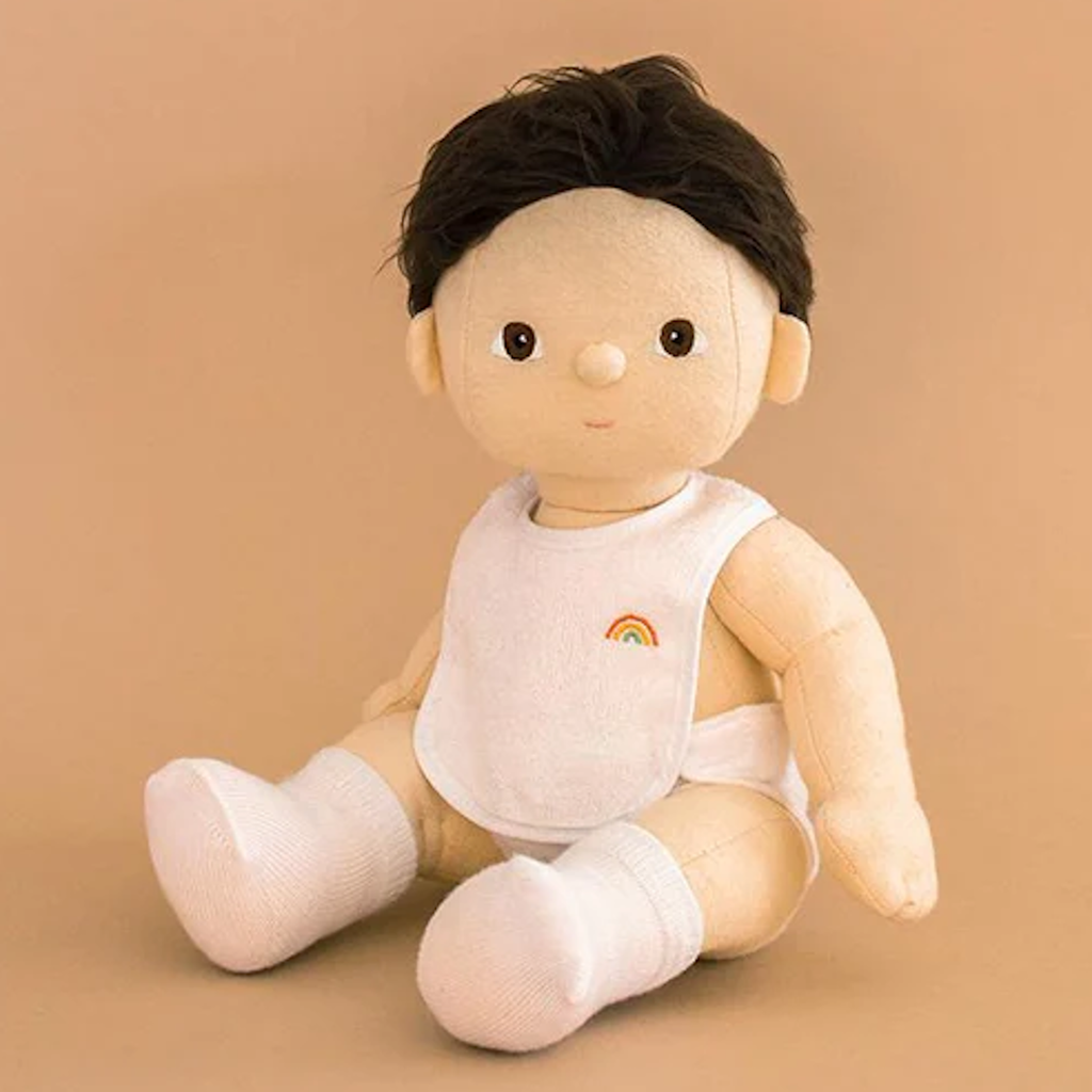 Plush doll with dark hair sits against a beige background, wearing a white bib with a rainbow, white socks, and a diaper. It has a soft, gentle expression.