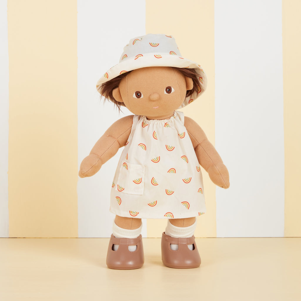 A soft fabric doll in a dress with rainbow patterns and a matching hat stands against a backdrop of vertical yellow and white stripes, wearing brown shoes.