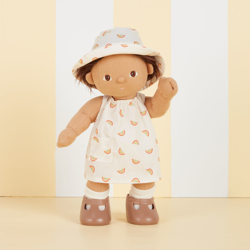 A soft fabric doll in a dress with rainbow patterns and a matching hat stands against a backdrop of vertical yellow and white stripes, wearing brown shoes.