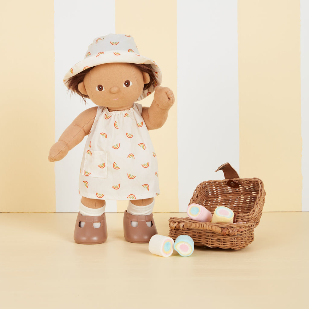 A soft fabric doll in a dress with rainbow patterns and a matching hat stands against a backdrop of vertical yellow and white stripes, wearing brown shoes.