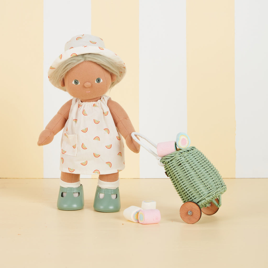 A soft fabric doll in a dress with rainbow patterns and a matching hat stands against a backdrop of vertical yellow and white stripes, wearing brown shoes.