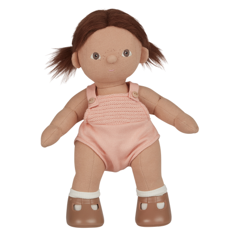 A soft, fabric doll with brown pigtails stands against a white background. It wears a pink romper and brown shoes, conveying a playful and charming tone.