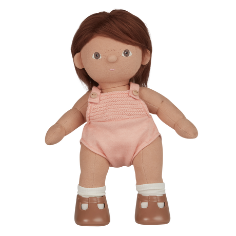 A soft, fabric doll with brown pigtails stands against a white background. It wears a pink romper and brown shoes, conveying a playful and charming tone.