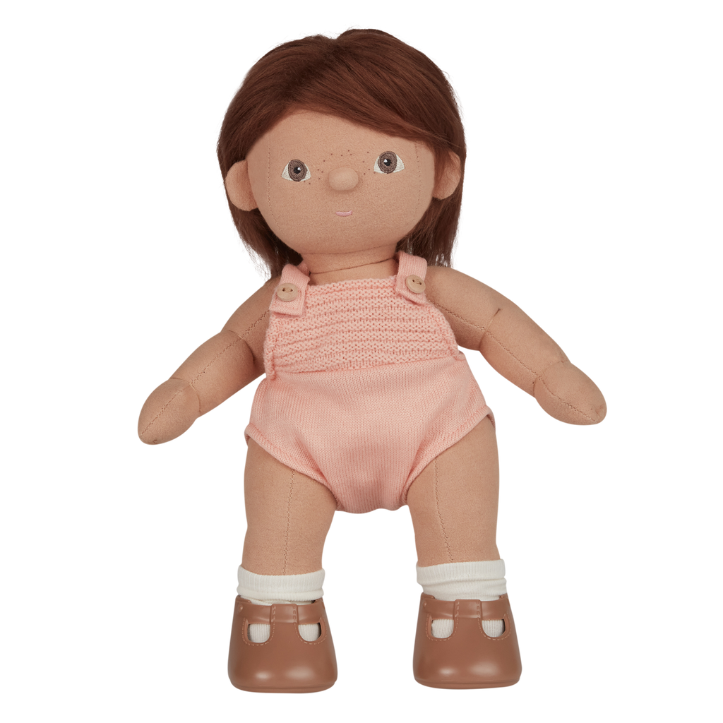 A soft, fabric doll with brown pigtails stands against a white background. It wears a pink romper and brown shoes, conveying a playful and charming tone.