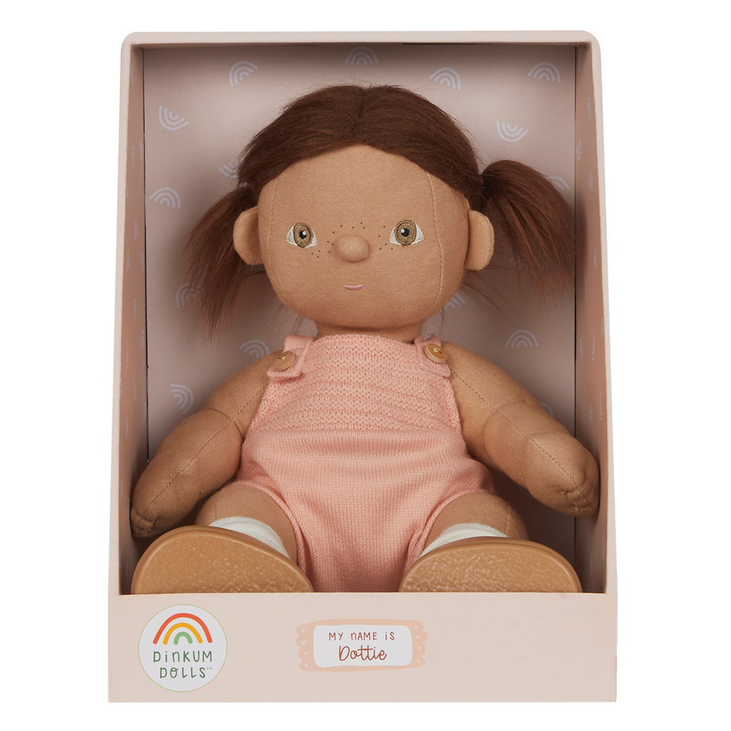 A soft, fabric doll with brown pigtails stands against a white background. It wears a pink romper and brown shoes, conveying a playful and charming tone.