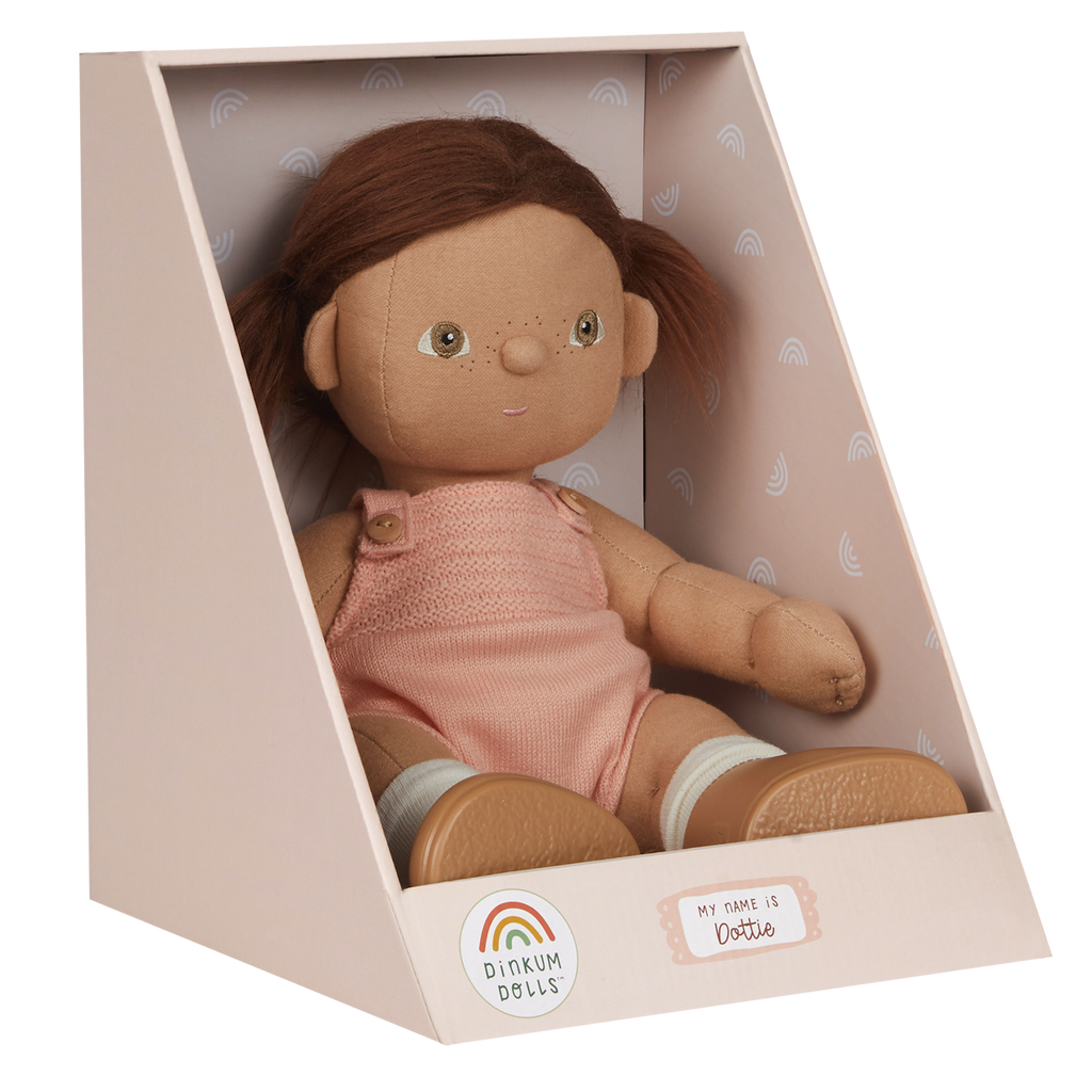 A soft, fabric doll with brown pigtails stands against a white background. It wears a pink romper and brown shoes, conveying a playful and charming tone.