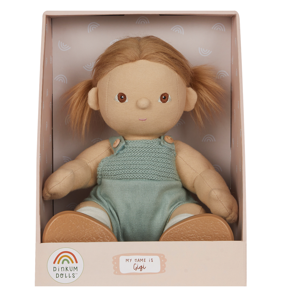 A soft doll with brown hair in pigtails sits in a pastel box. The doll wears a knitted green romper and is labeled "Gigi" with a cheerful expression.