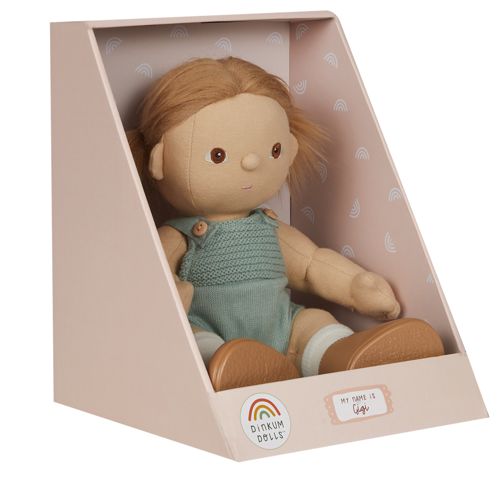 A soft doll with brown hair in pigtails sits in a pastel box. The doll wears a knitted green romper and is labeled "Gigi" with a cheerful expression.