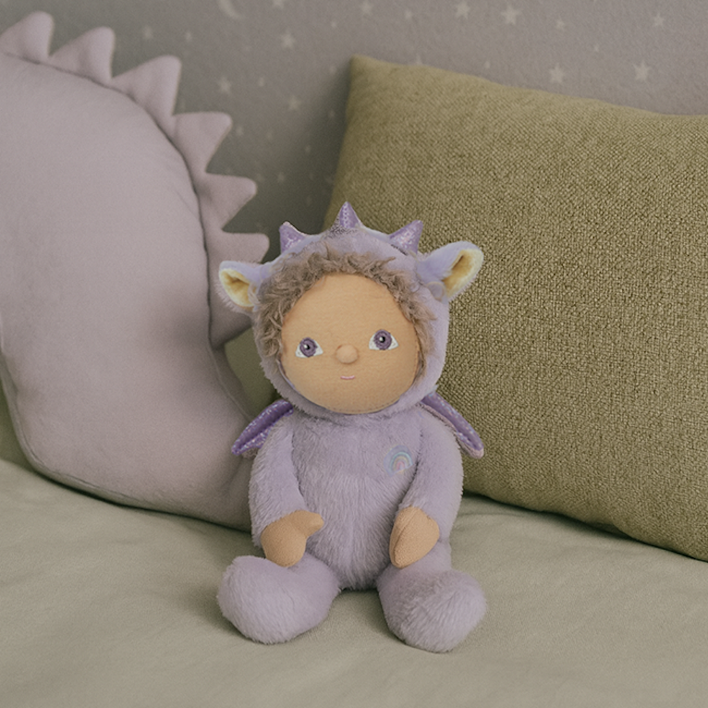 A plush doll wearing a lavender dragon costume with sparkly ears and horns. The doll has curly hair, a serene expression, and a rainbow emblem.