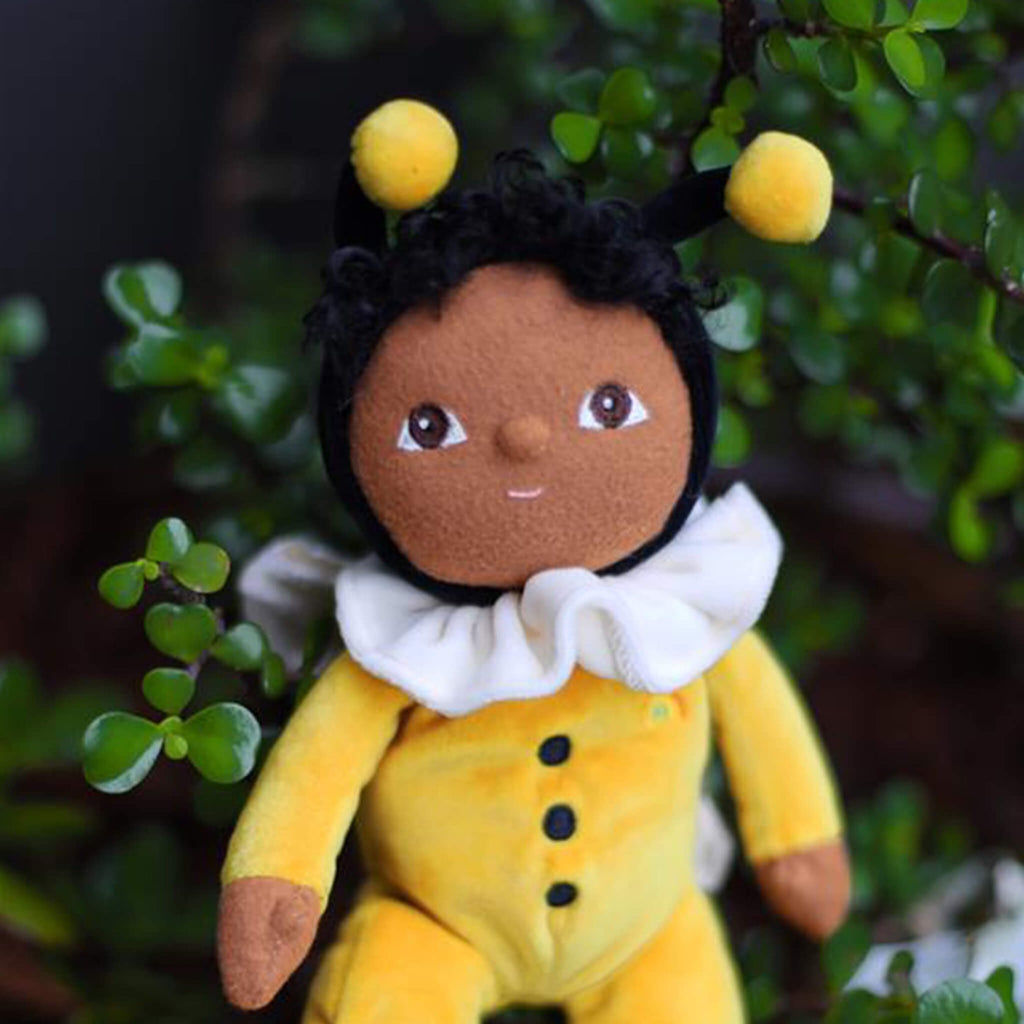 Plush doll dressed as a bee, featuring a black head with bee antennae, a white ruffled collar, and yellow outfit with wing details.