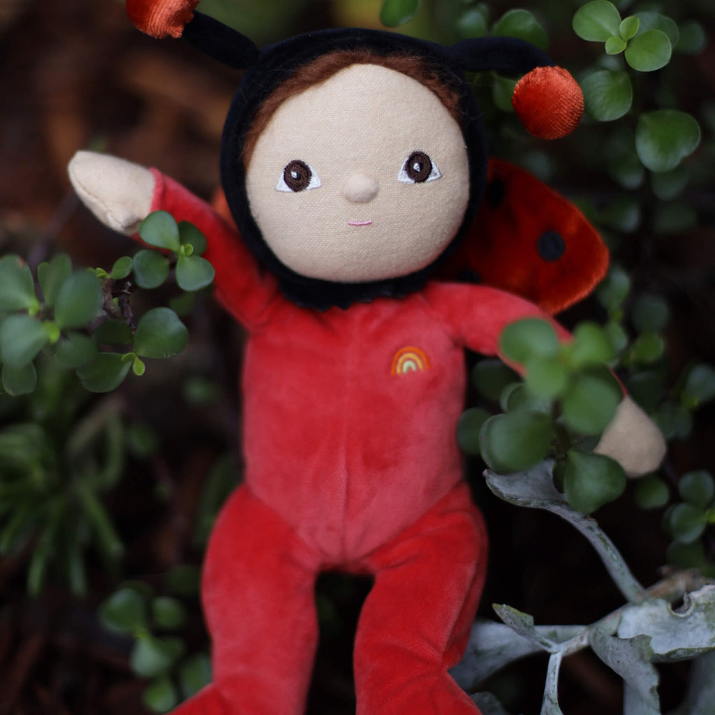 Soft plush toy resembling a ladybug figure, wearing a red outfit with a rainbow patch. Features a gentle facial expression and antennae on its hood.