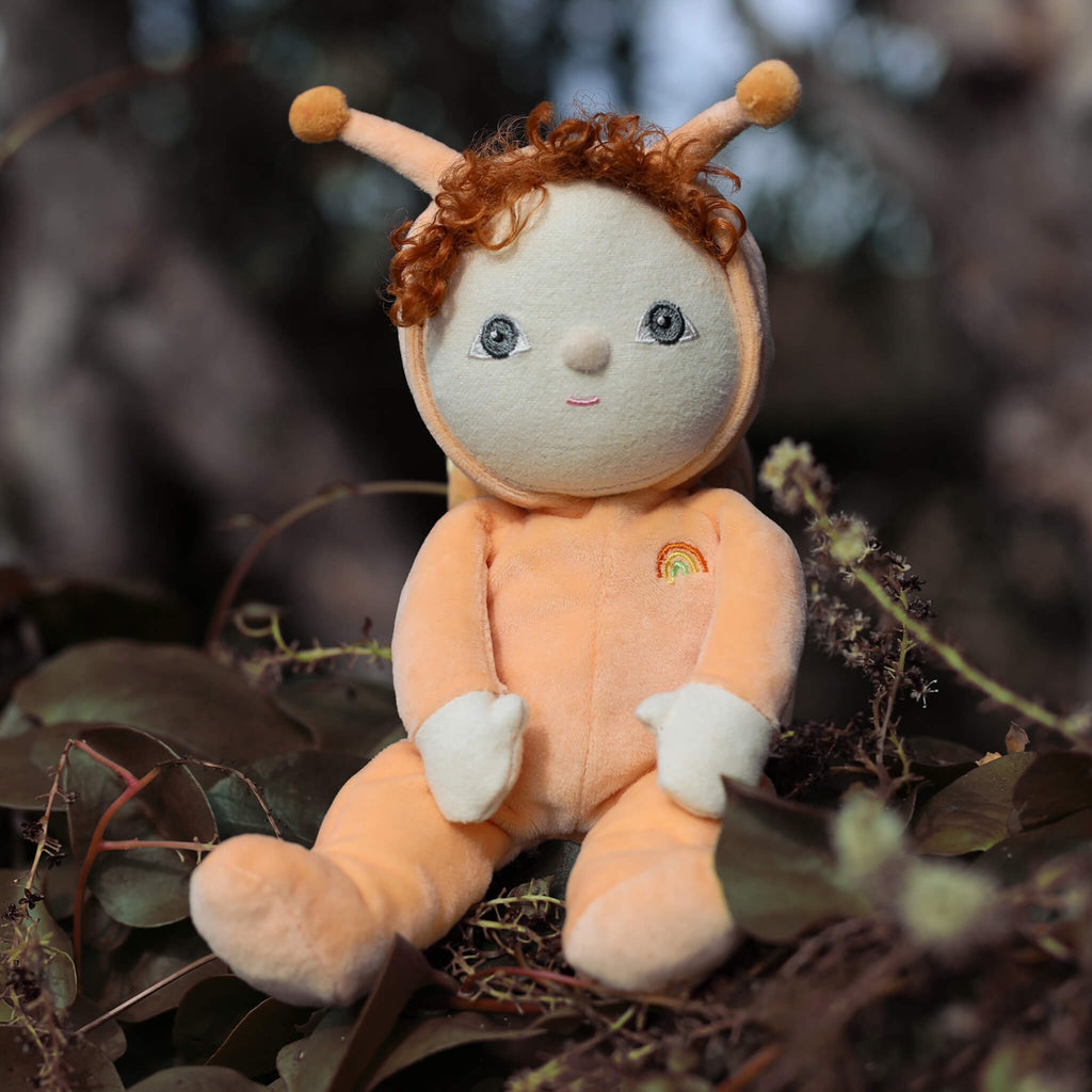 Cute plush doll with curly red hair, dressed in a peach snail outfit with antennas and a rainbow emblem. It sits with a sweet, cheerful expression.