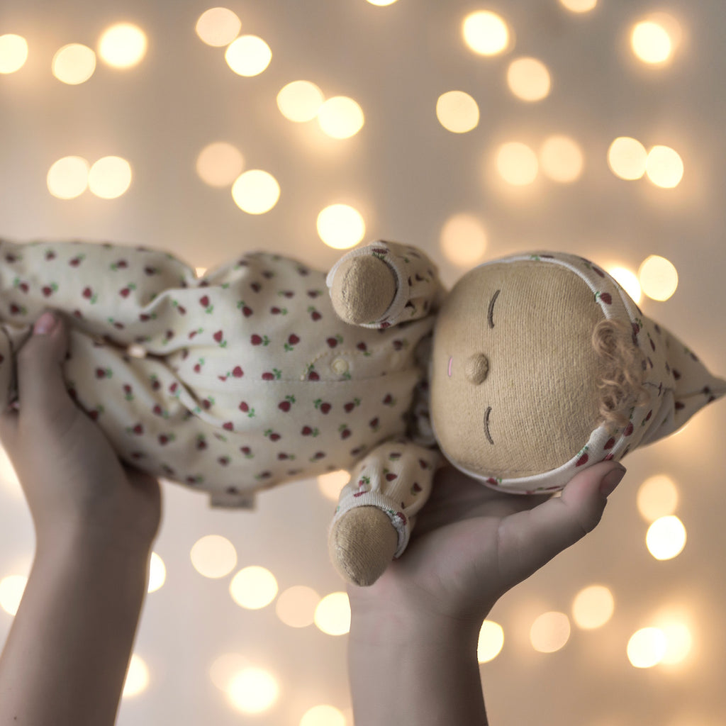 A soft doll in a cream-colored outfit with red strawberry patterns. The doll has closed eyes, curly hair on top, and small mittens and booties.