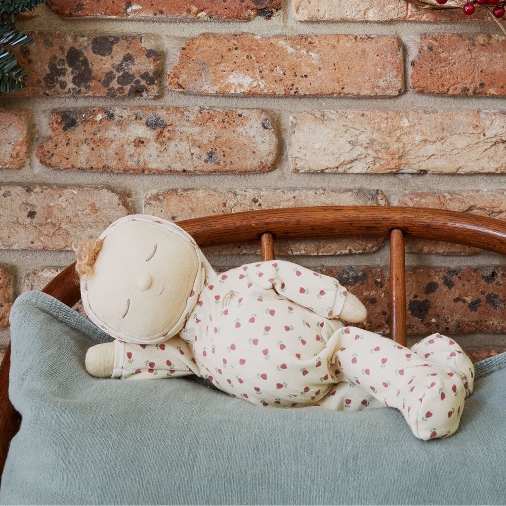 A soft doll in a cream-colored outfit with red strawberry patterns. The doll has closed eyes, curly hair on top, and small mittens and booties.
