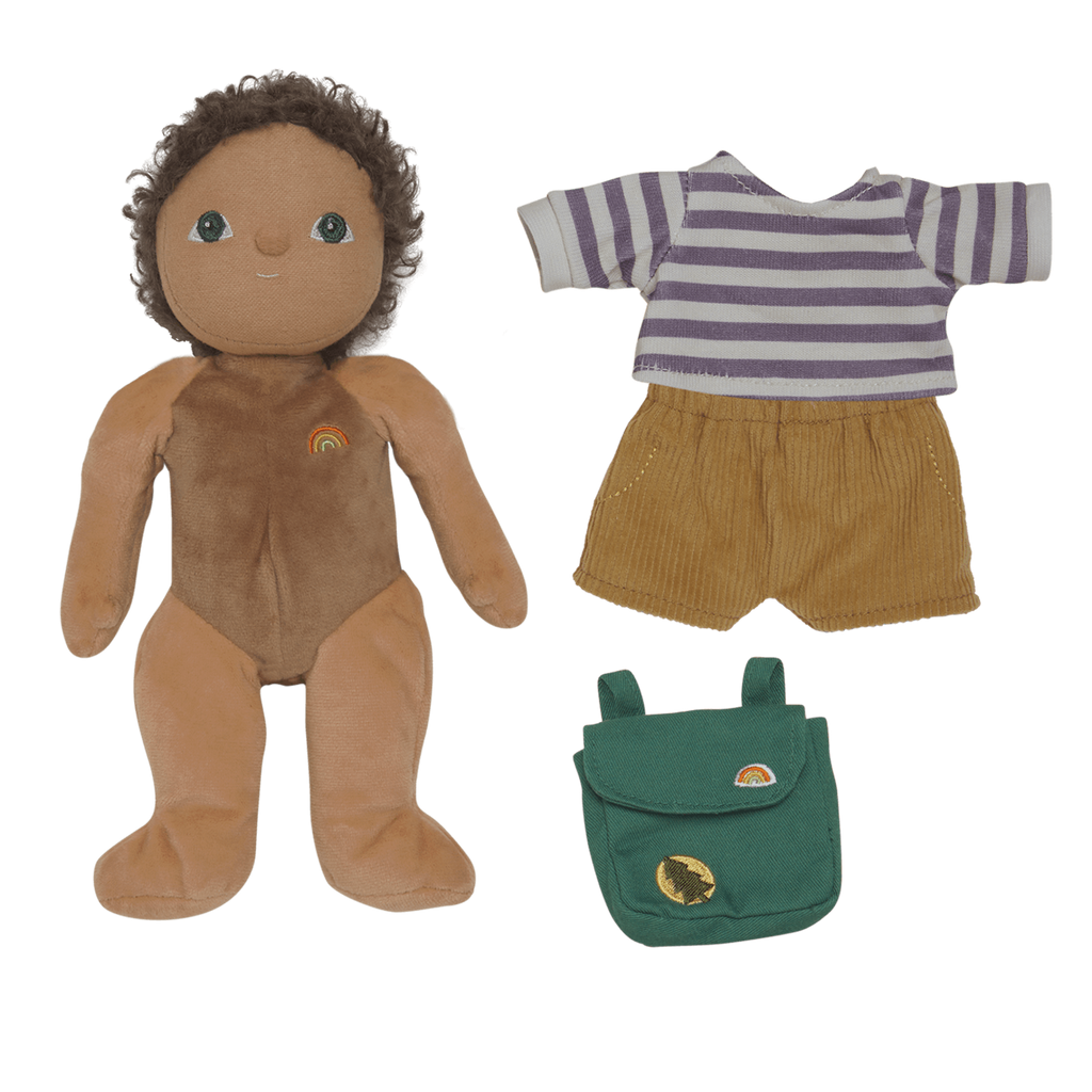 A plush doll with curly hair and a rainbow emblem on its chest lies next to a striped shirt, tan shorts, and a small green backpack on a white background.