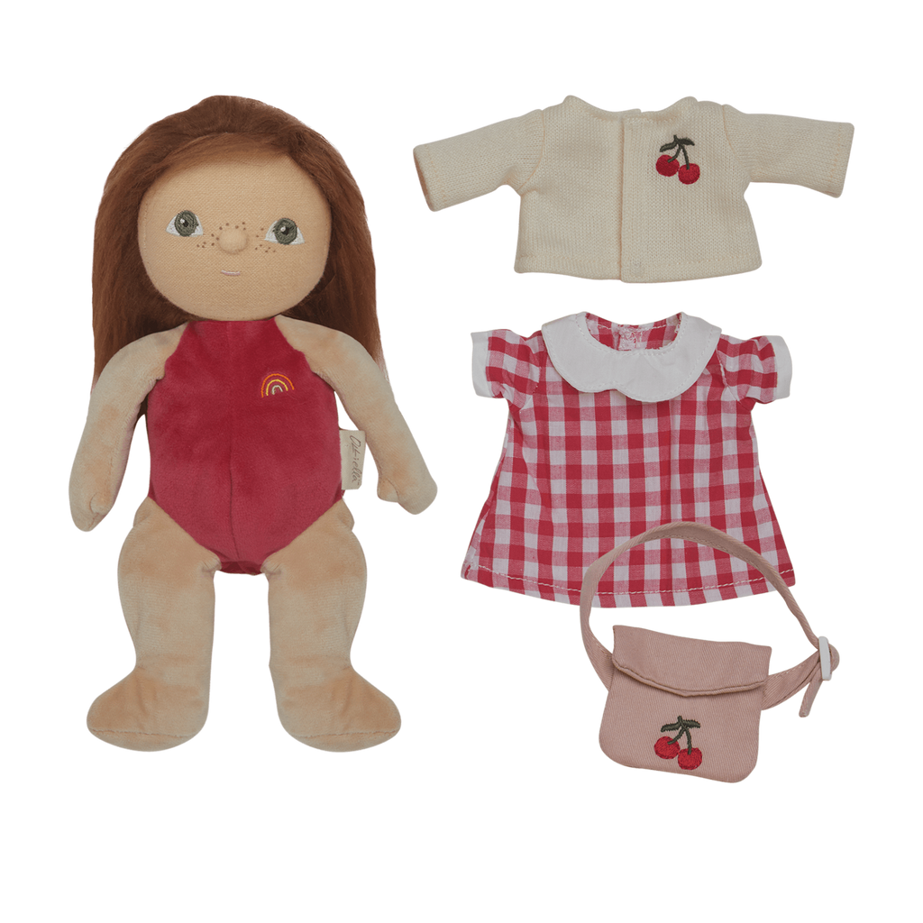 A soft doll with brown hair in a red outfit lies next to clothes: a cherry-embroidered cream cardigan, a red checkered dress, and a matching cherry bag.
