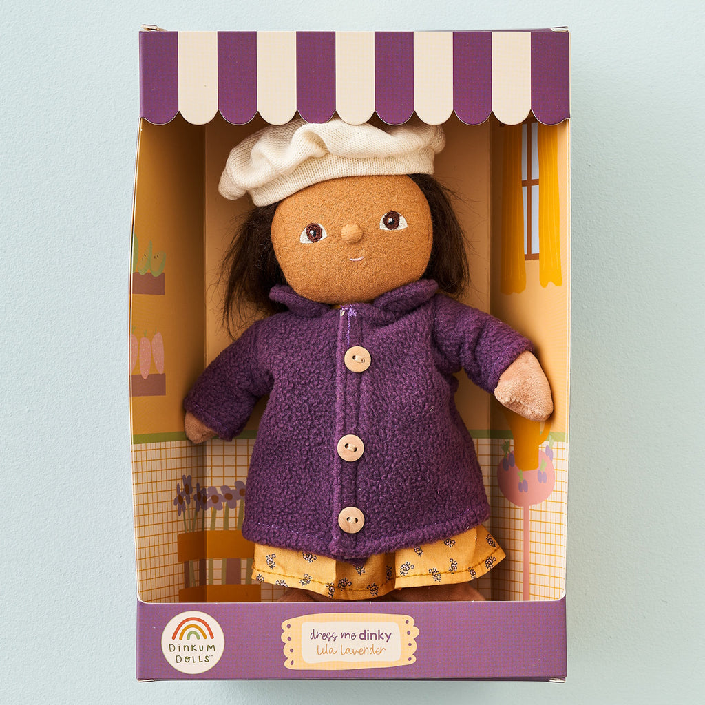 A plush doll with brown hair, wearing a purple coat and white chef's hat, is displayed in a decorative box with purple and white striped edges.