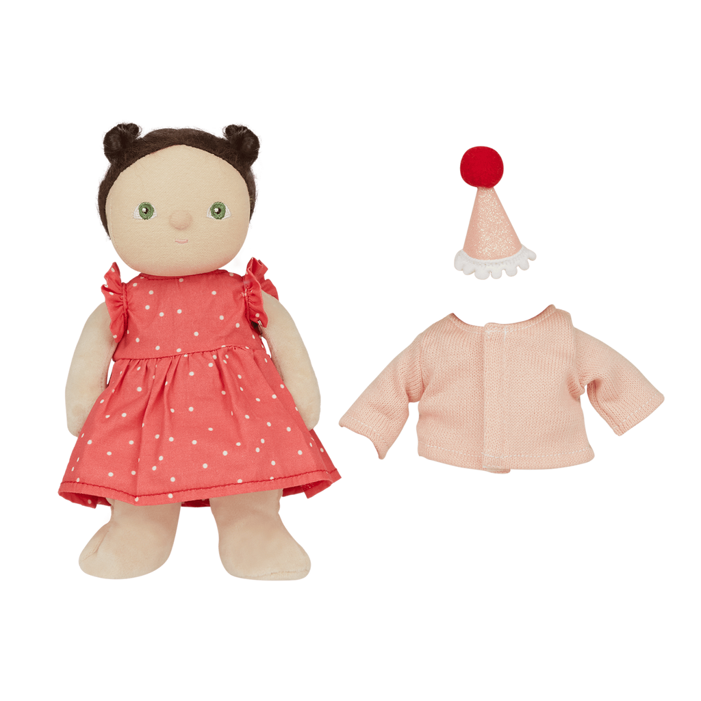 A soft doll in a red polka dot dress with brown hair buns. Nearby are a pink cardigan and a pink party hat with a red pompom, against a white background.