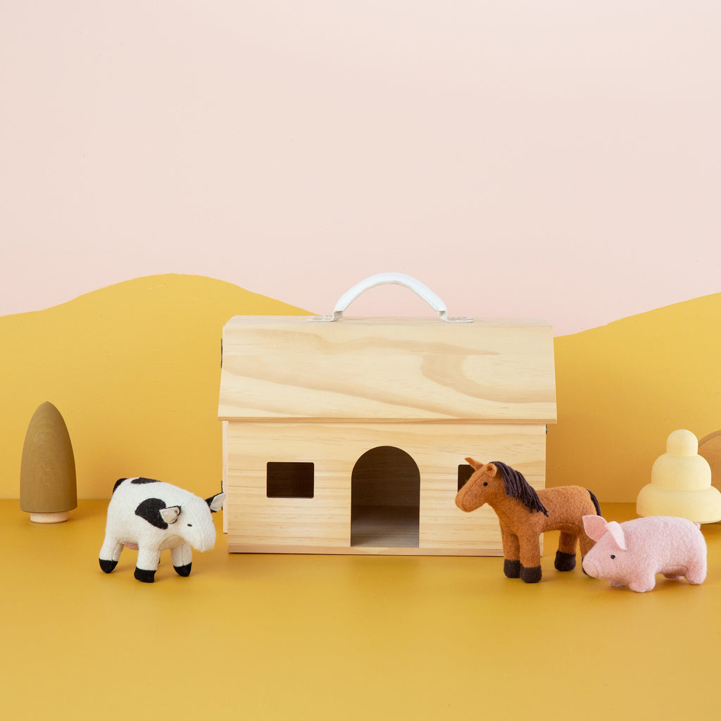 Wooden toy barn with felt animals, including a black and white cow, brown horse, and pink pig, on a pastel yellow and pink background. Playful and whimsical.