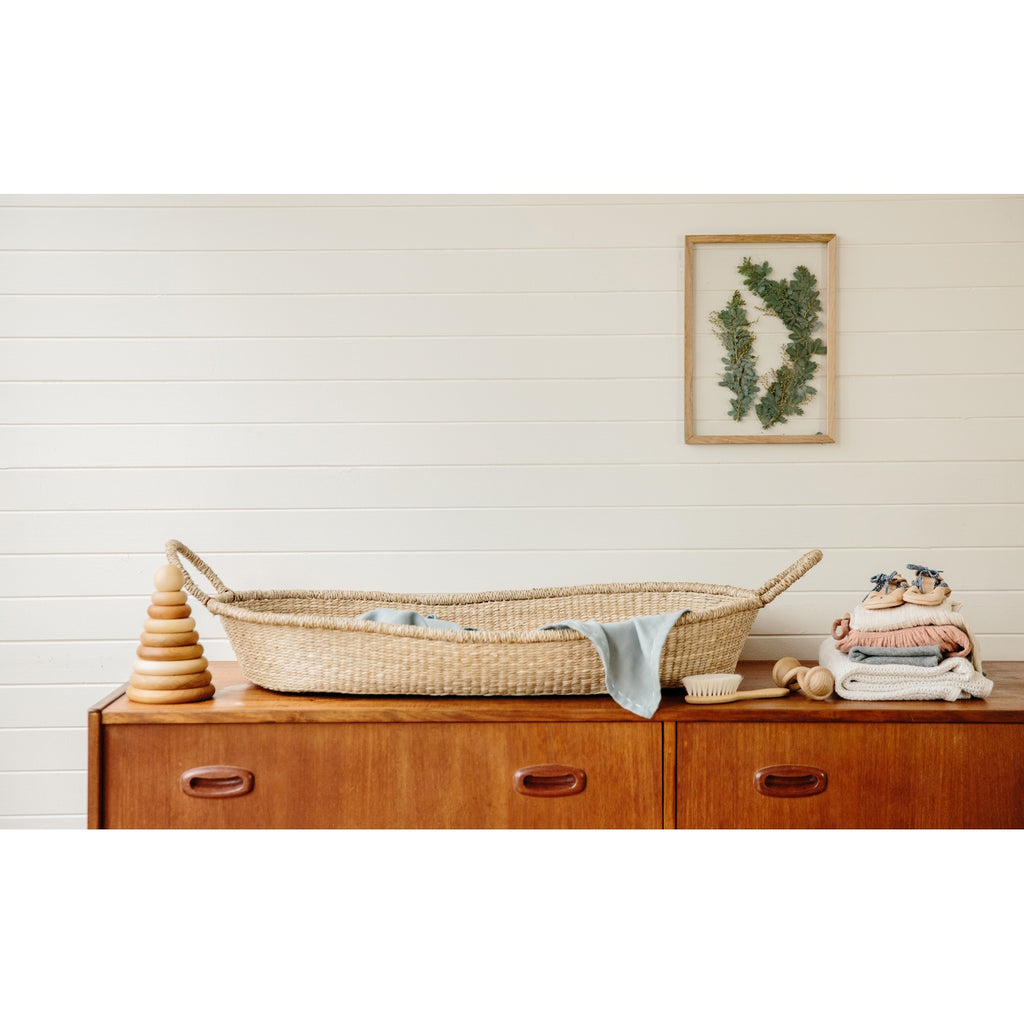 A natural baby changing basket with two loop handles on each side. The basket is oval-shaped, with a simple, rustic texture and a neutral, earthy tone.