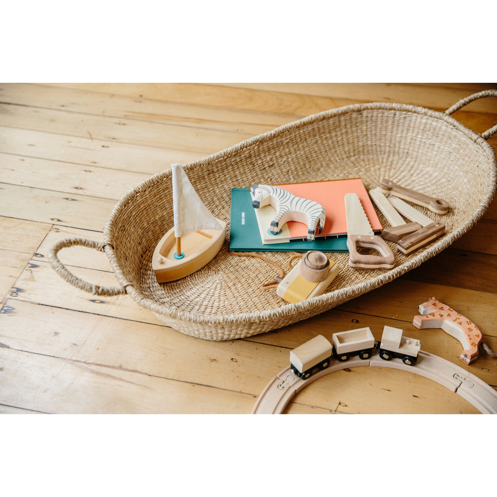 A natural baby changing basket with two loop handles on each side. The basket is oval-shaped, with a simple, rustic texture and a neutral, earthy tone.