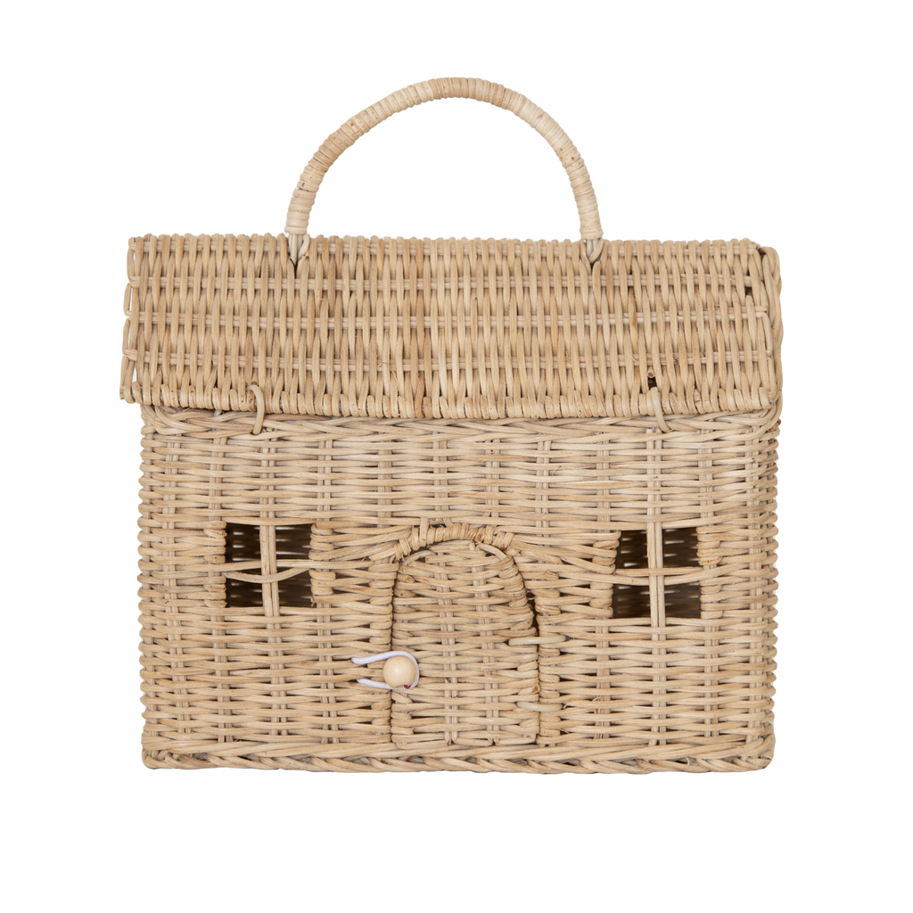 Wicker basket shaped like a small house, featuring a woven handle, arched door with button clasp, and window patterns on the front. Neutral tones on white background.