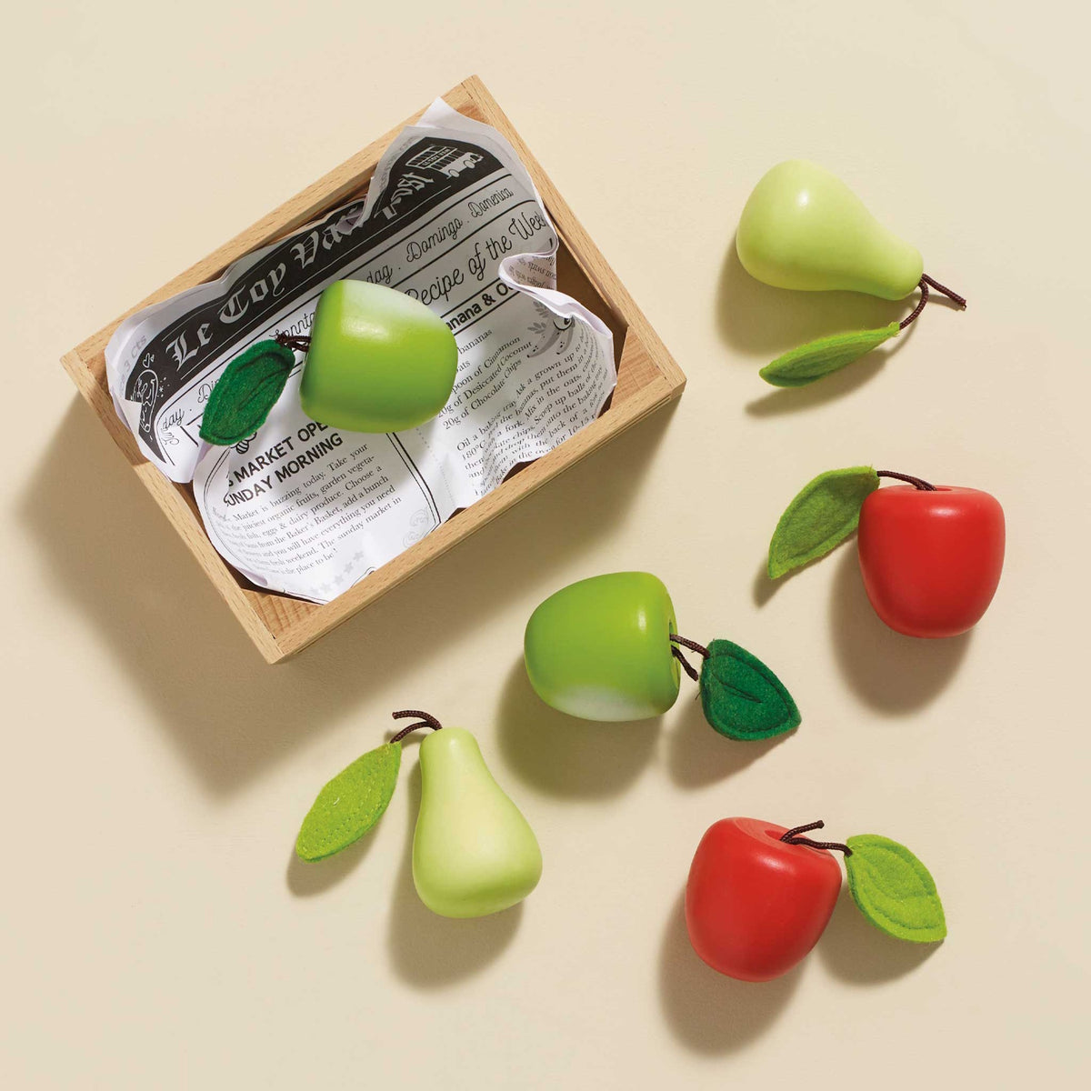 Wooden toy crate and scattered apples and pears with felt leaves on a beige background.