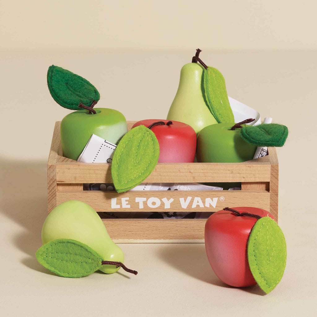 Wooden crate labeled "Le Toy Van" filled with brightly colored toy apples and pears, some red, some green. Felt leaves add a playful touch.