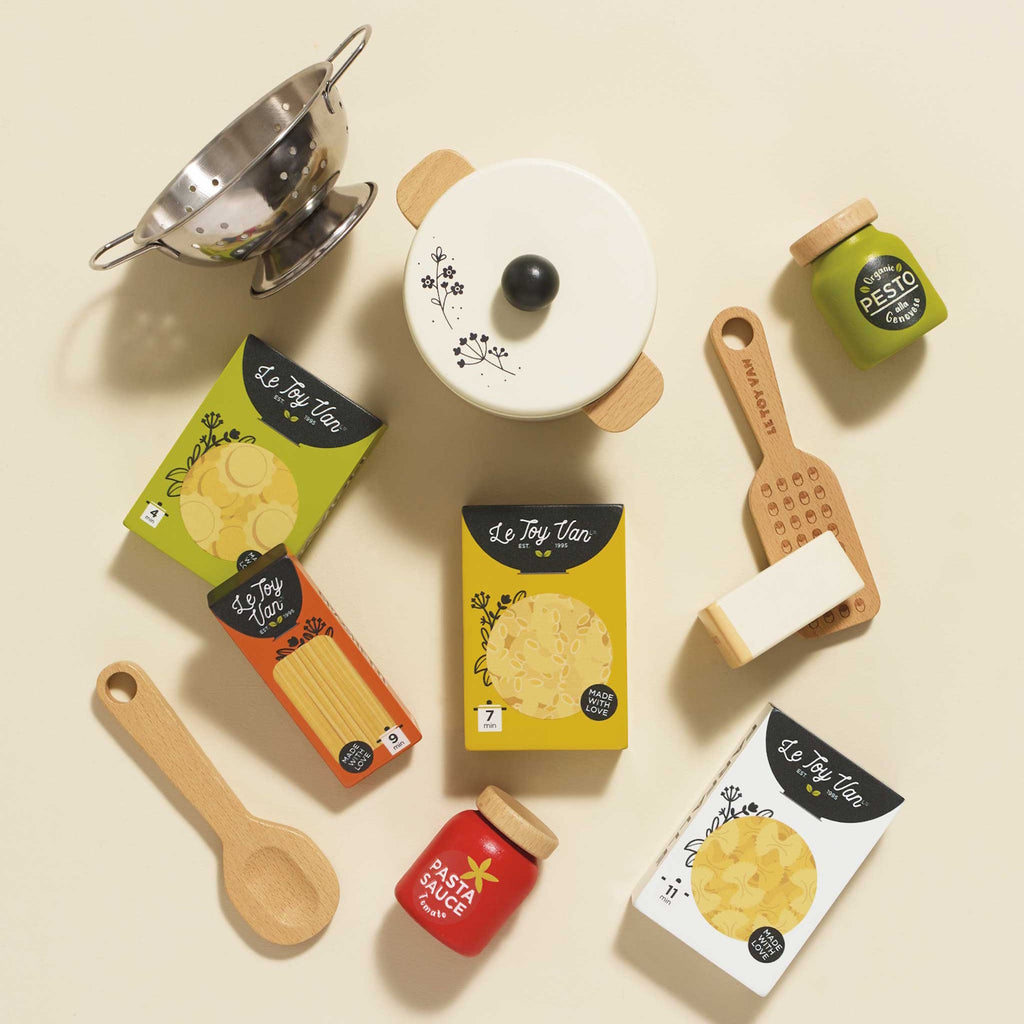 Toy pasta set featuring a cream pot with a lid, wooden spoon, cheese wedge, colander, pesto, and pasta sauce jars, alongside boxed pasta shapes.