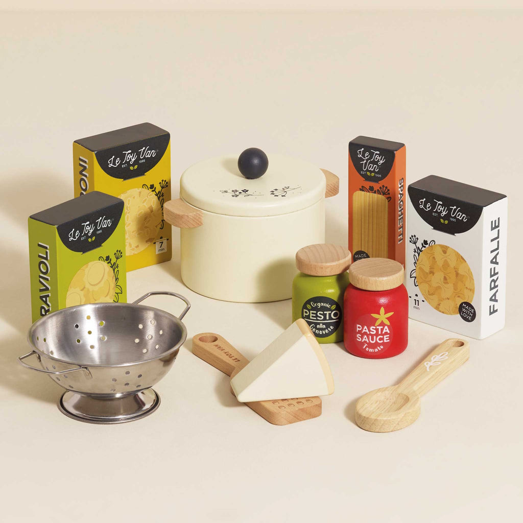 Toy pasta set featuring a cream pot with a lid, wooden spoon, cheese wedge, colander, pesto, and pasta sauce jars, alongside boxed pasta shapes.