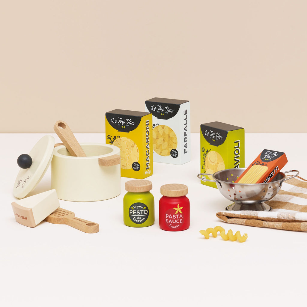Toy pasta set featuring a cream pot with a lid, wooden spoon, cheese wedge, colander, pesto, and pasta sauce jars, alongside boxed pasta shapes.
