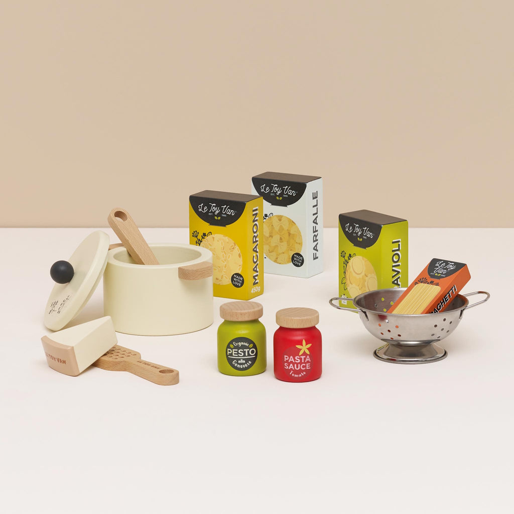 Toy pasta set featuring a cream pot with a lid, wooden spoon, cheese wedge, colander, pesto, and pasta sauce jars, alongside boxed pasta shapes.