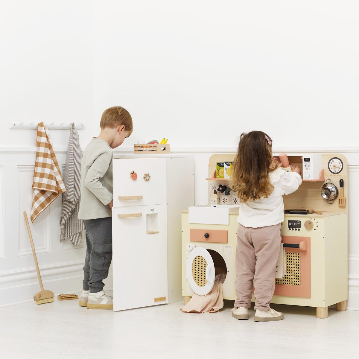 Two young children playing with a wooden toy fridge and play kitchen set in a white room.