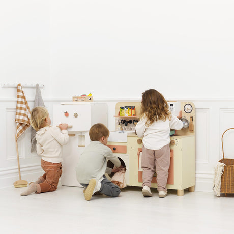 Three young children playing together with a wooden toy fridge and play kitchen set in a bright room.