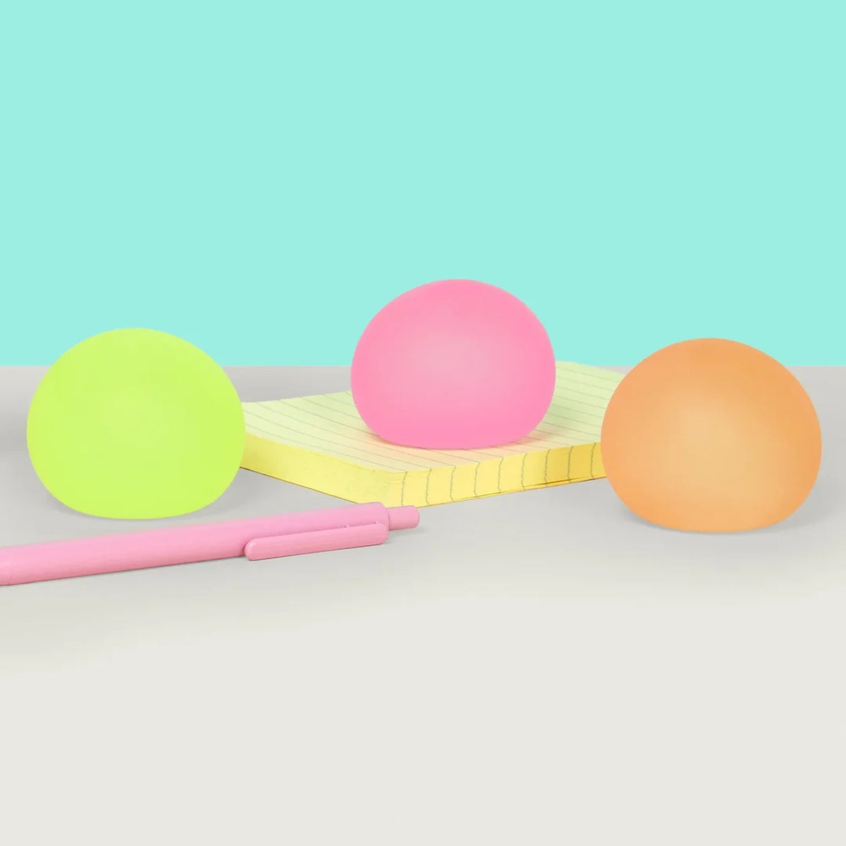Three colorful memory gel stress balls sitting on a desk with a yellow notepad and pink pen.