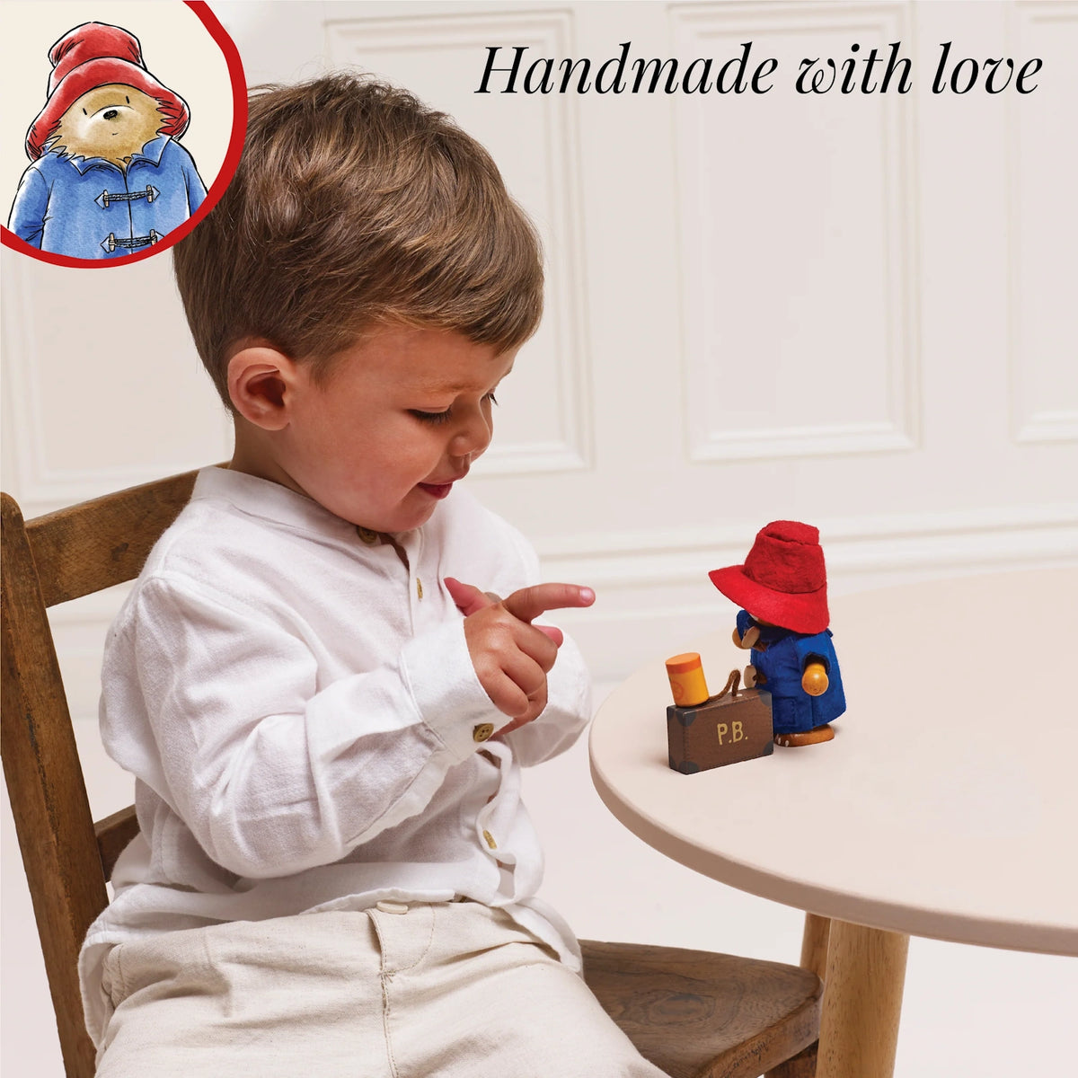 Young boy sitting at a wooden table pointing at a Paddington Bear toy figure with text "Handmade with love".
