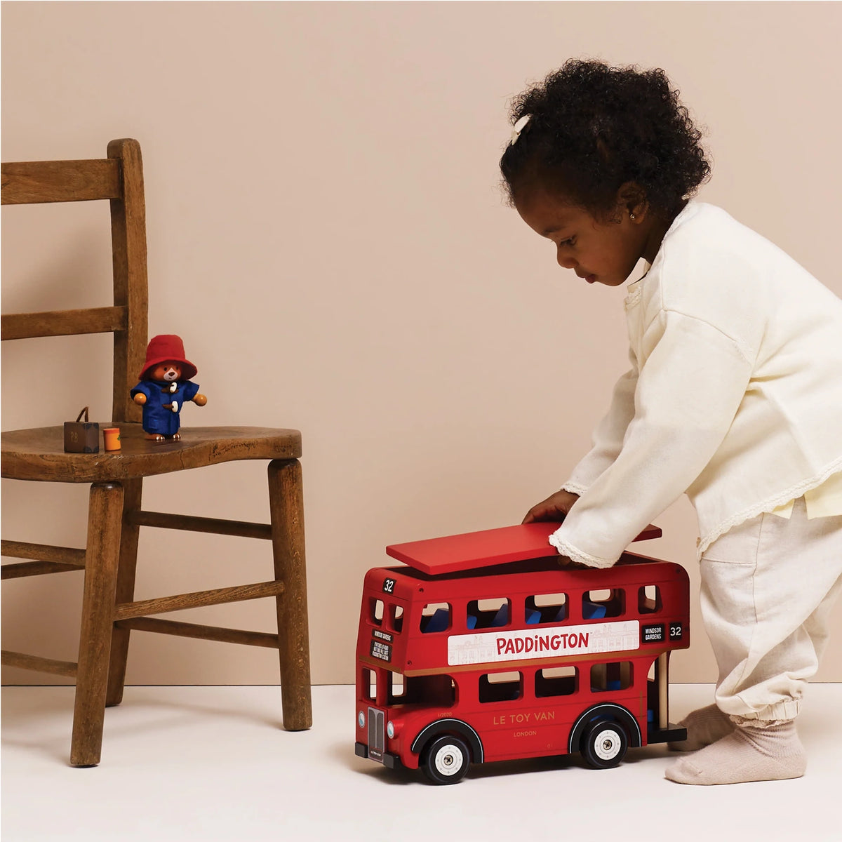 A young child playing with a wooden red London double-decker bus toy, with a toy Paddington Bear on a chair nearby.