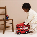 A young child playing with a wooden red London double-decker bus toy, with a toy Paddington Bear on a chair nearby.